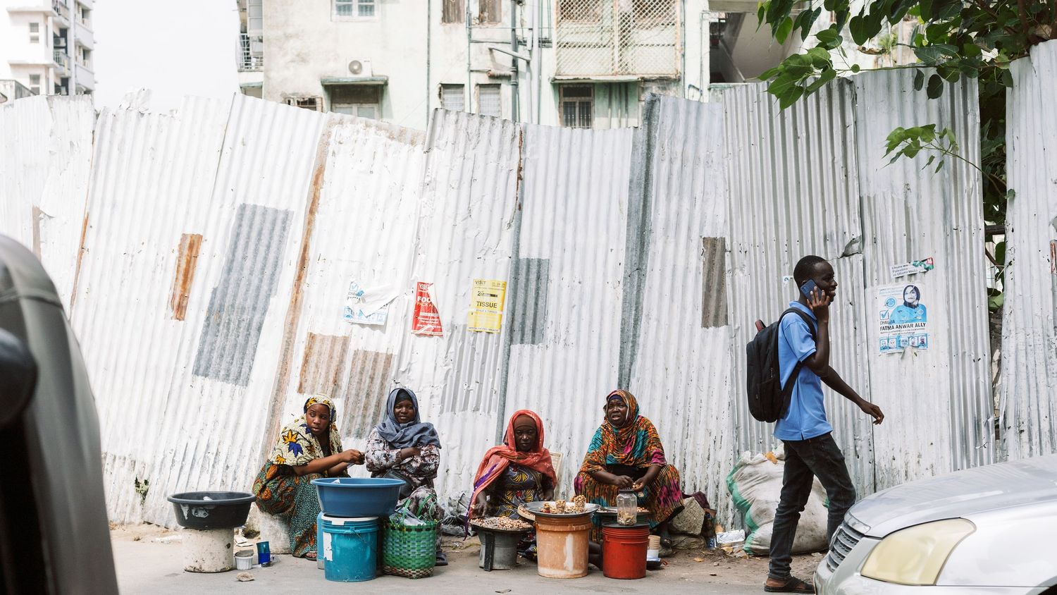 Women selling bites and talking near a corrugated iron fence, authentic scene of daily social life in Dar es Salaam streets.