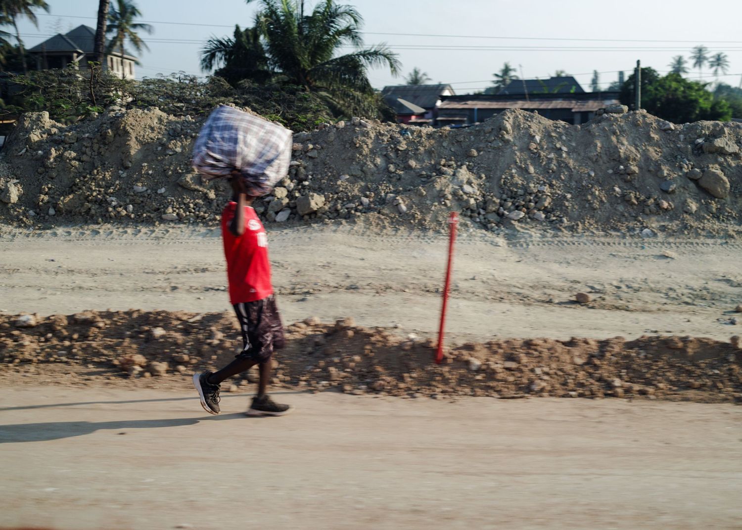 Man carrying a heavy checkered sack on his head walking past a construction site, capturing the stride and resilience of Tanzanian labor.