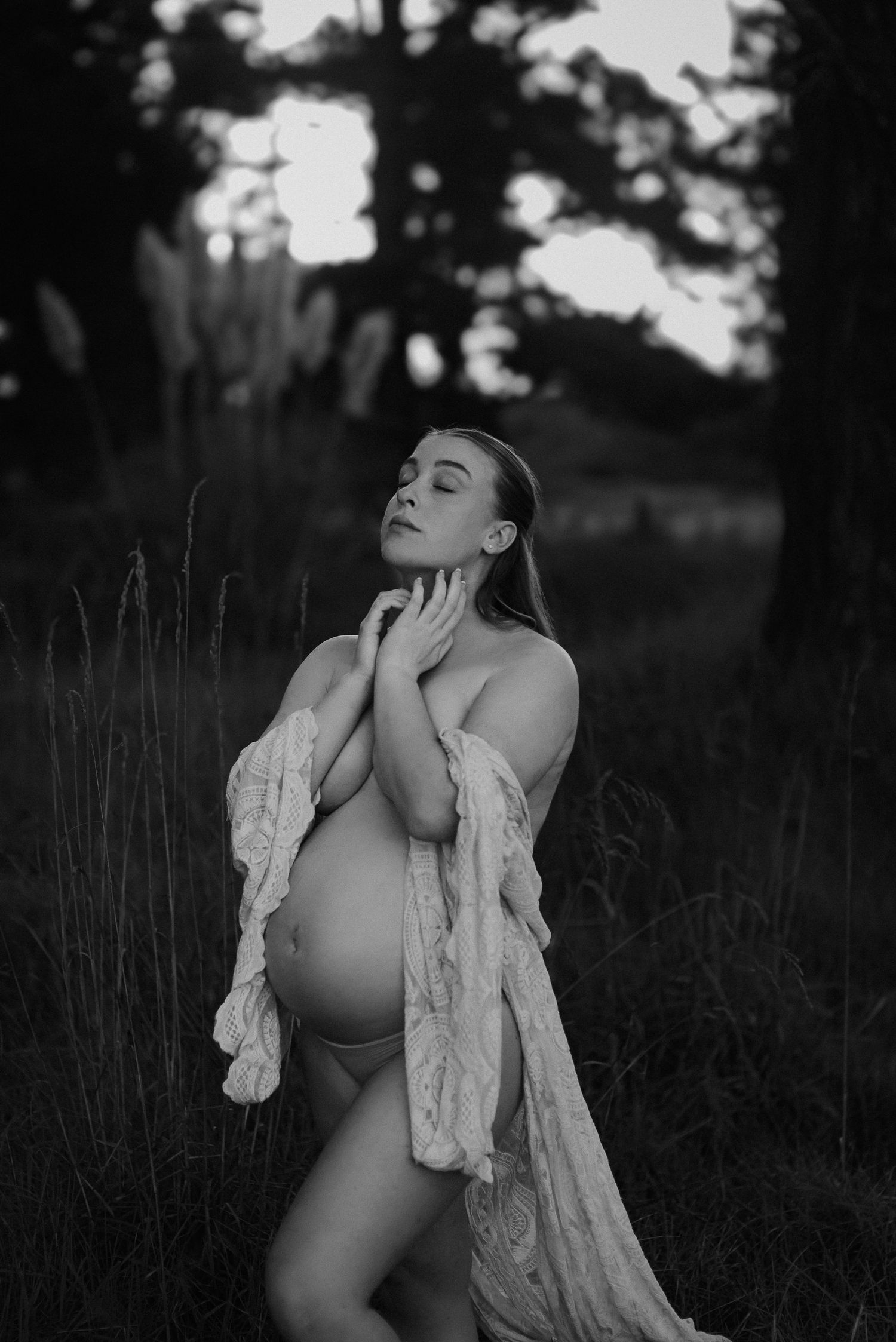 Artistic maternity photo in black and white showing an ethereal pose in a field at dusk.