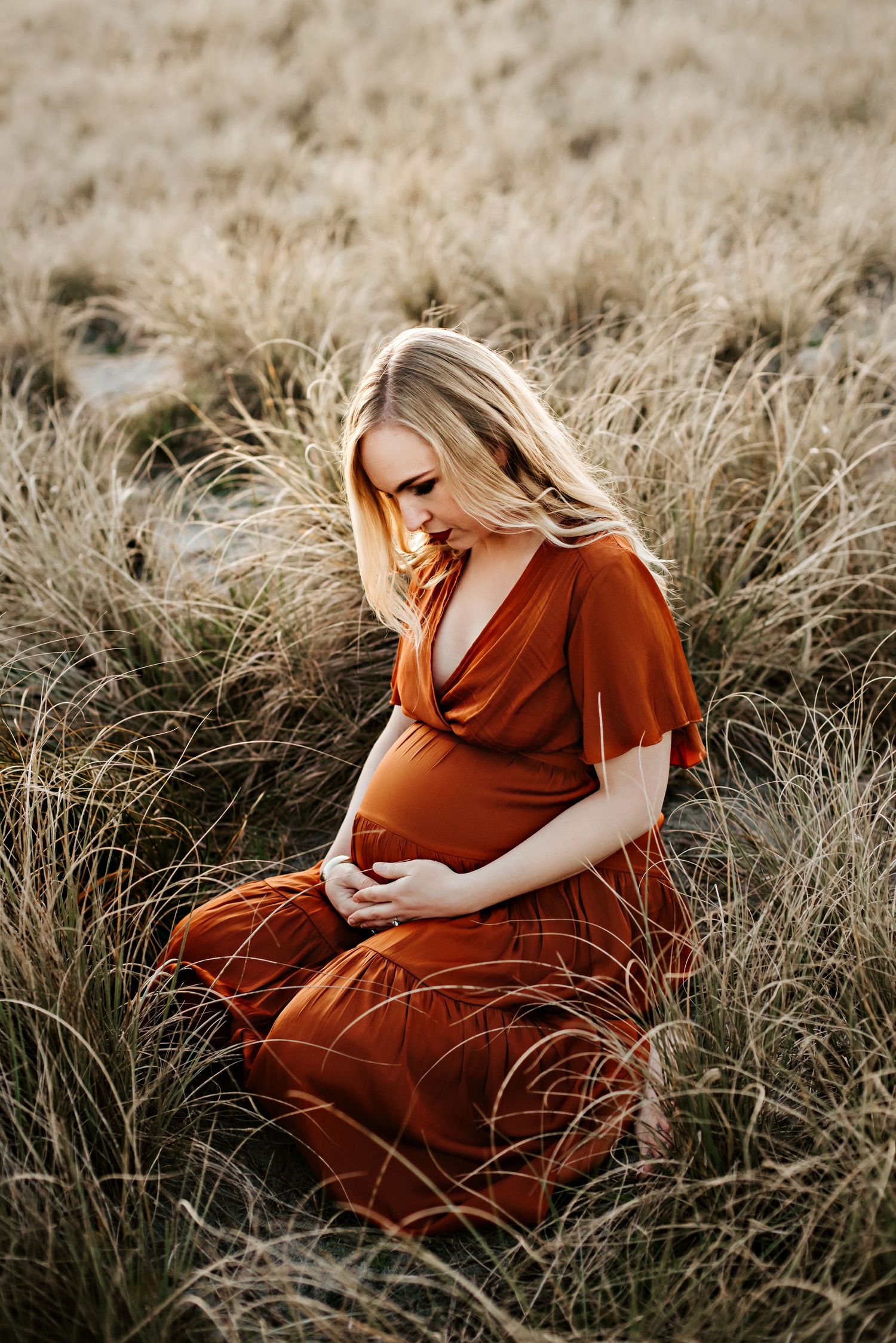 Maternity portrait in rust-colored dress sitting among tall golden grass at sunset.