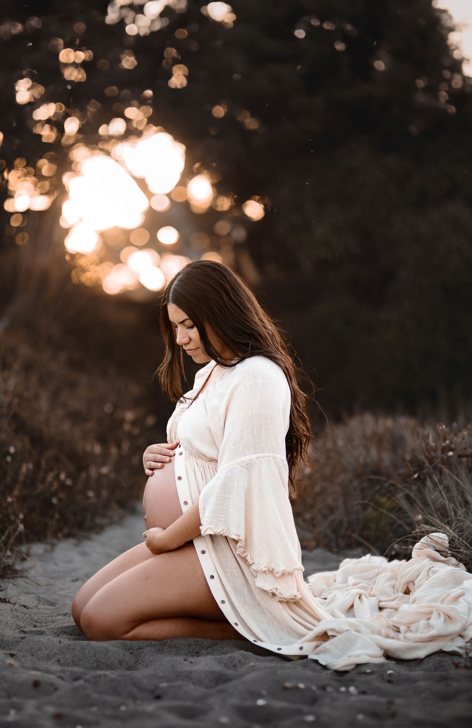 Maternity photo of an expectant mother in white dress sitting on beach at sunset with bokeh lighting.