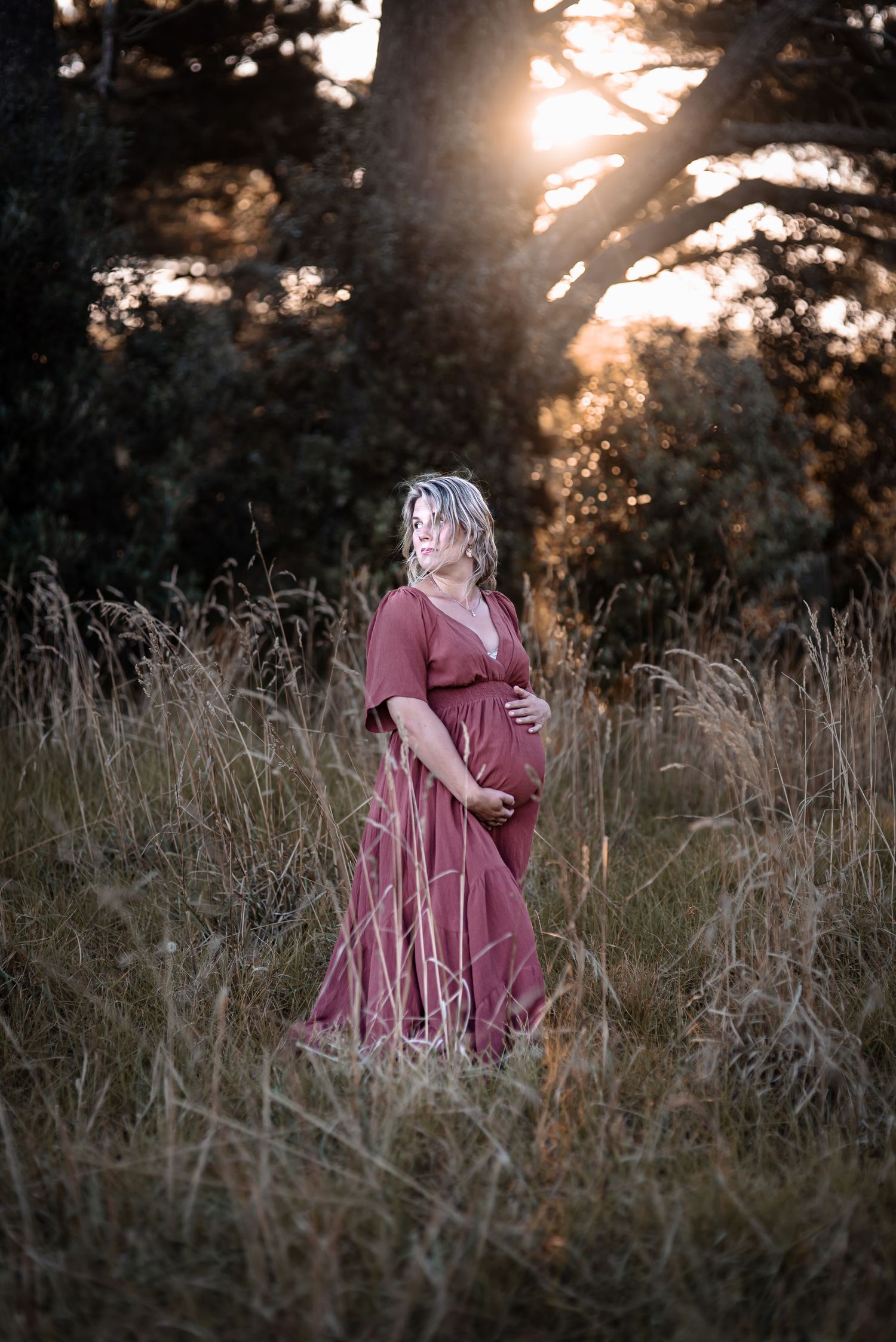 A maternity photo featuring a woman in a burgundy dress at sunset in a field.
