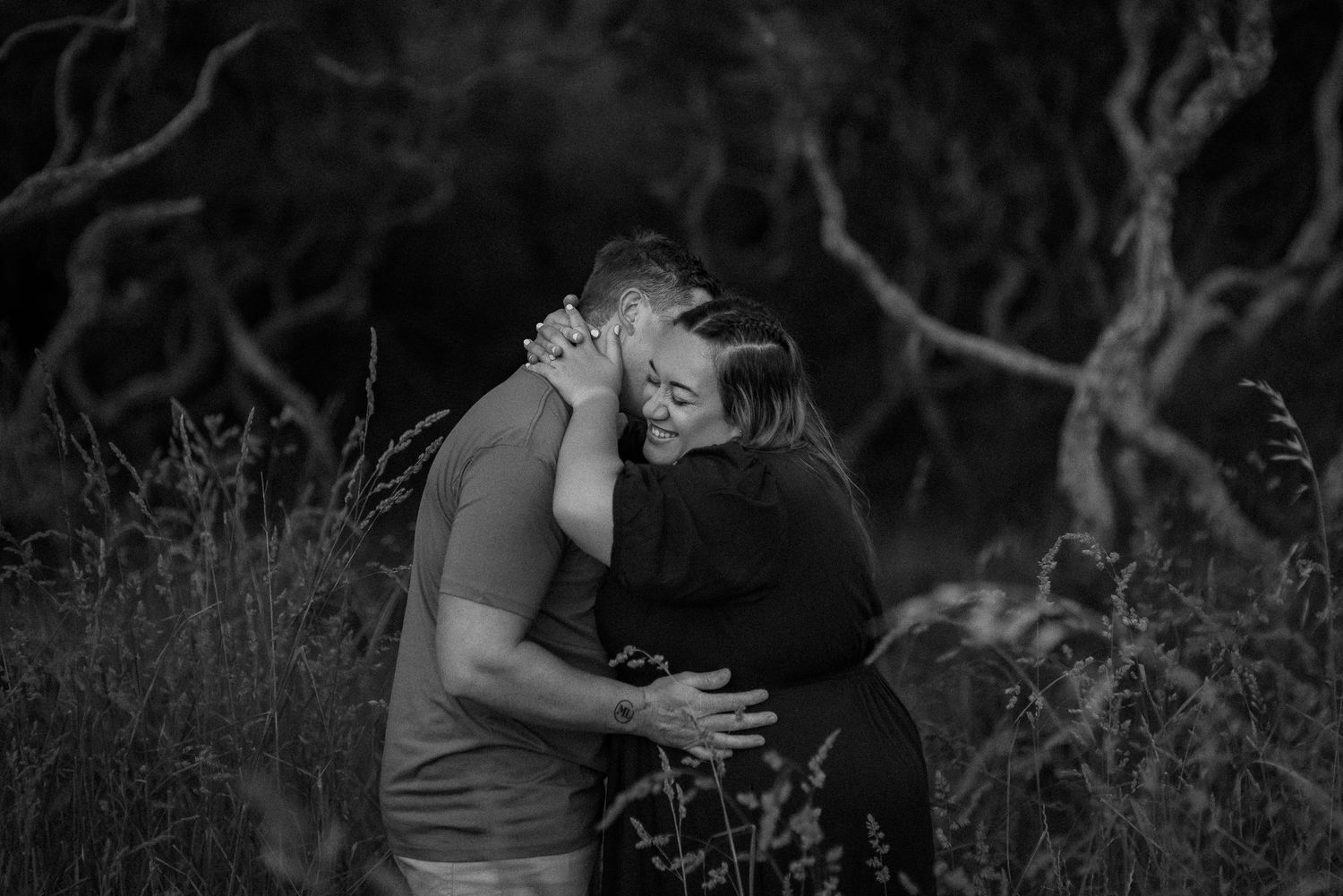 An intimate black and white portrait of a couple embracing outdoors among trees.
