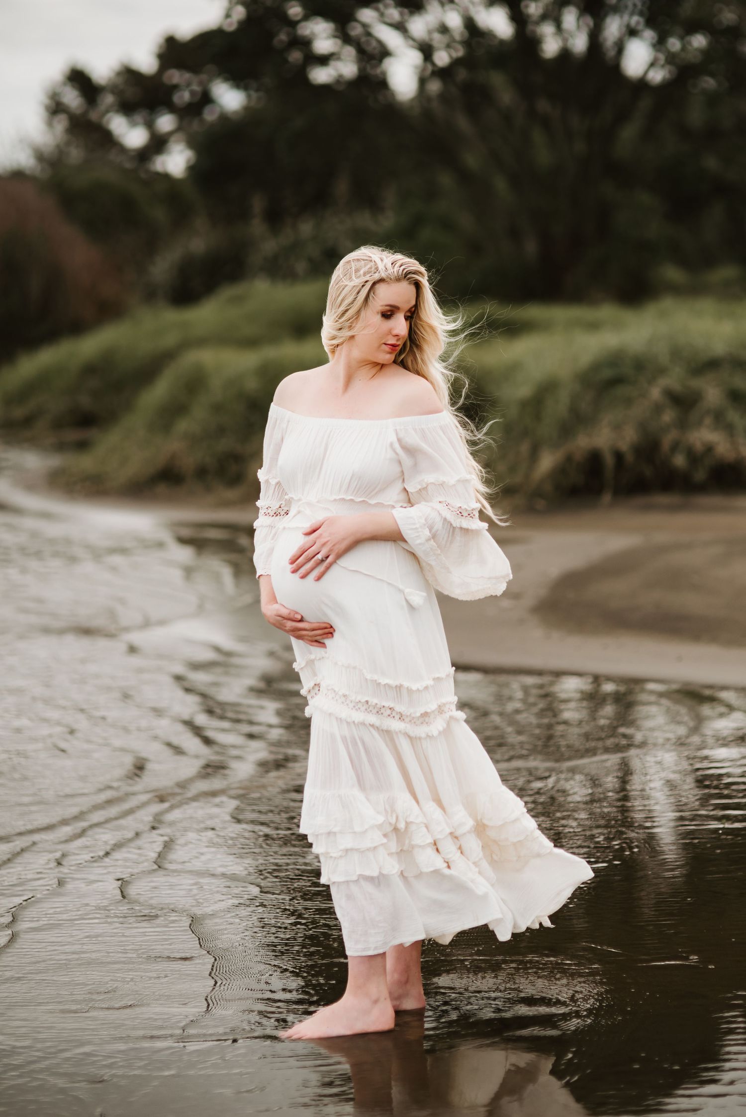 A maternity photo of a woman in a white ruffled dress standing in shallow water.