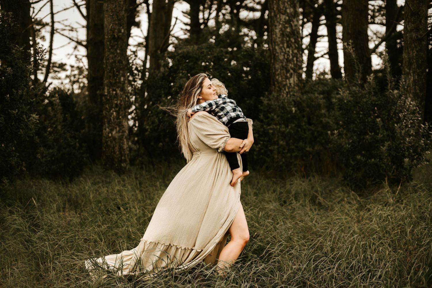 Artistic maternity photo in flowing beige dress surrounded by tall grass and trees.