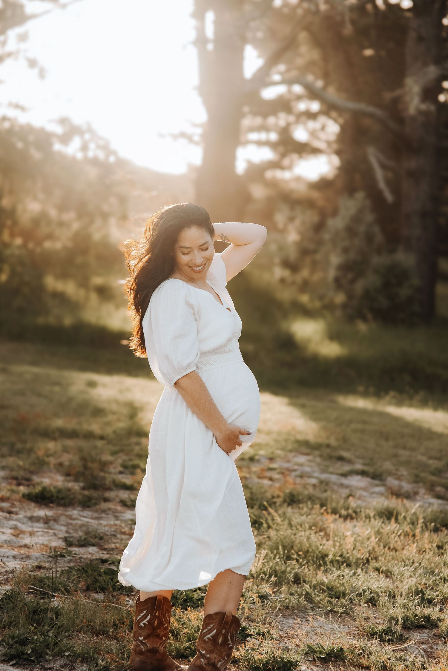 Sunset maternity photo of woman in white dress and boots in golden outdoor setting.