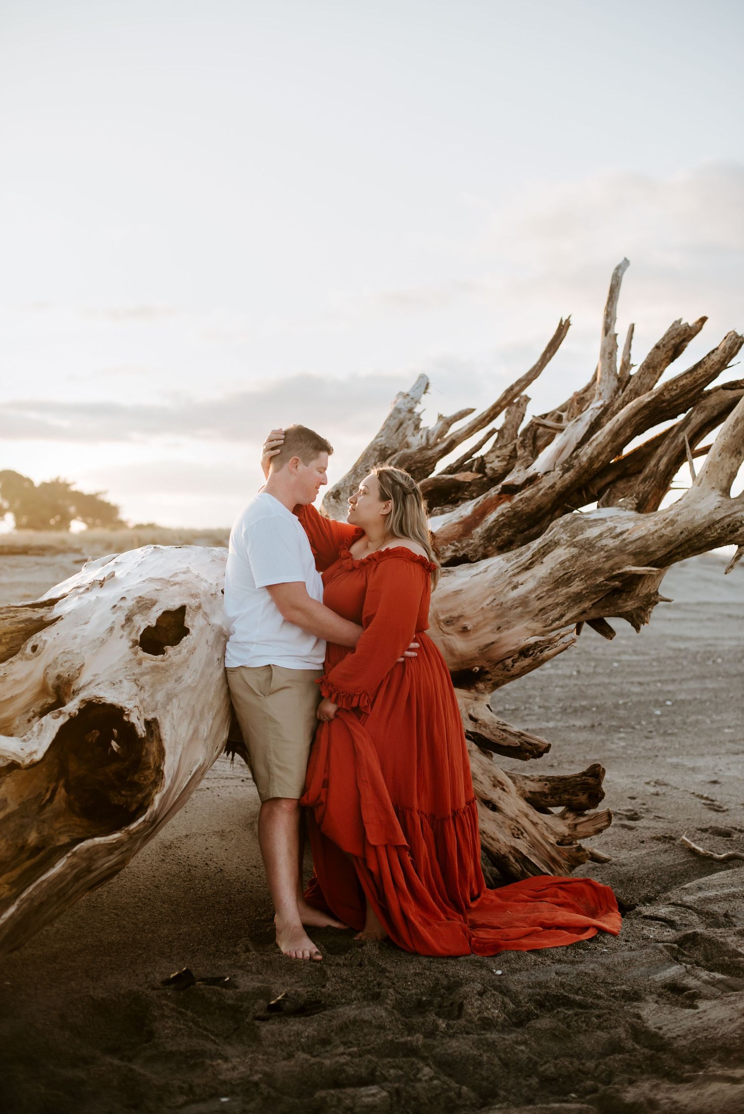 Romantic sunset beach photoshoot with couple embracing near driftwood on sandy shore.