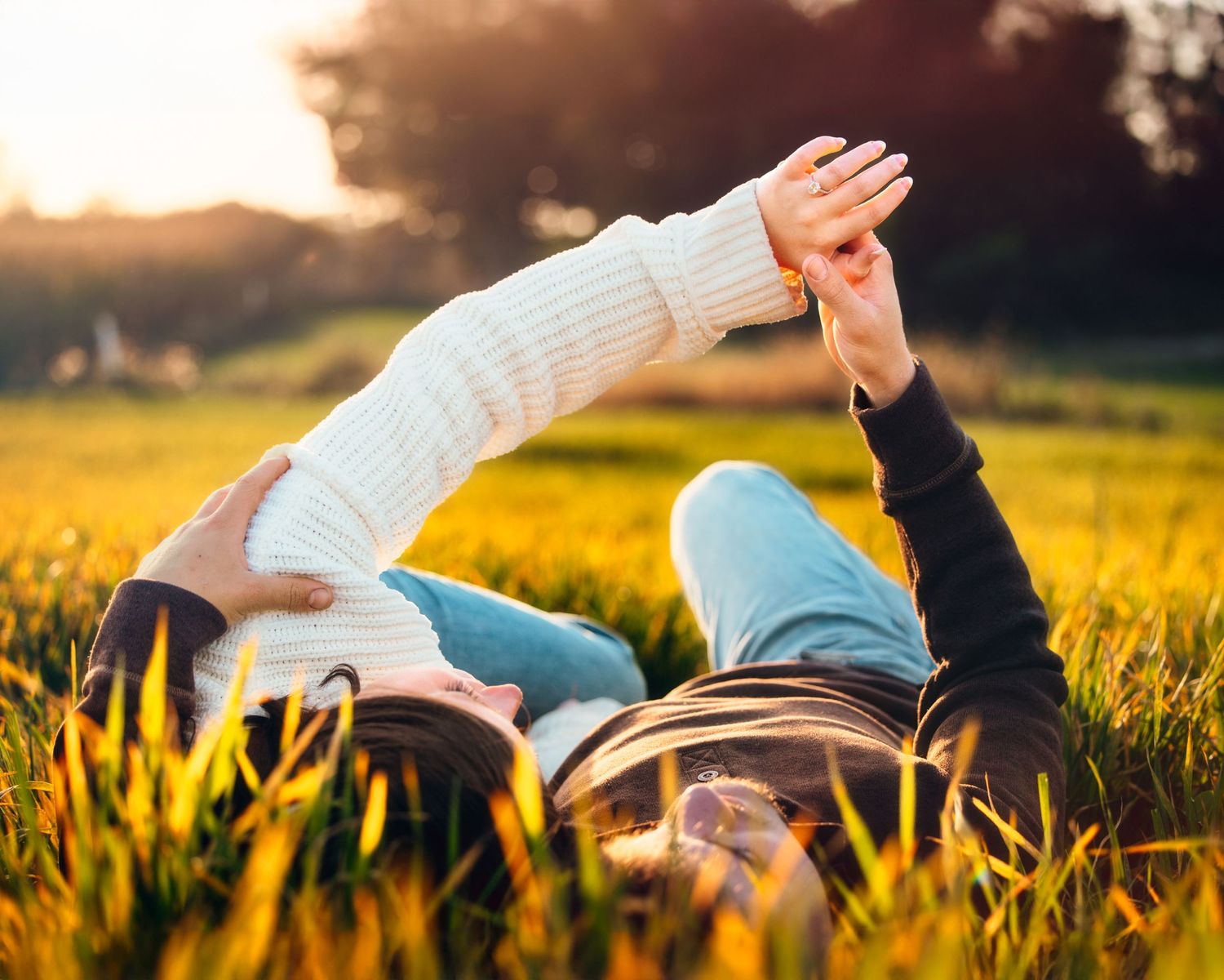 Couple lying in grass field during golden hour with arms outstretched.