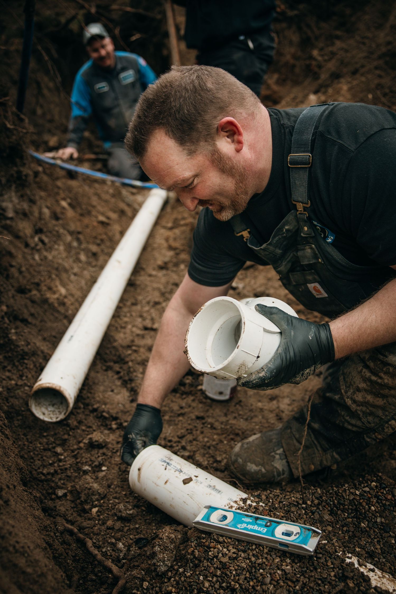 Plumber installing white PVC pipe in excavated trench.