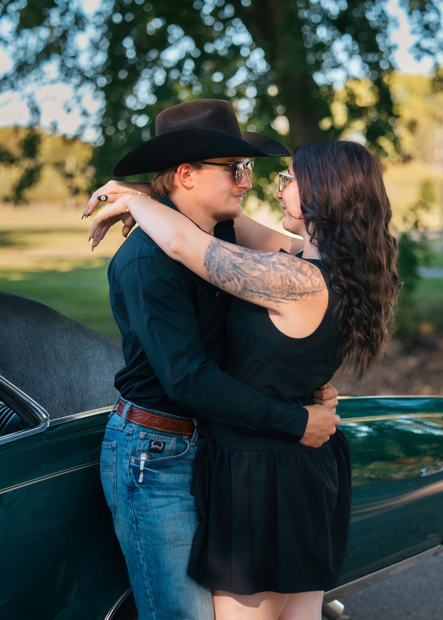 Couple embraces near vintage car with green trees in background.