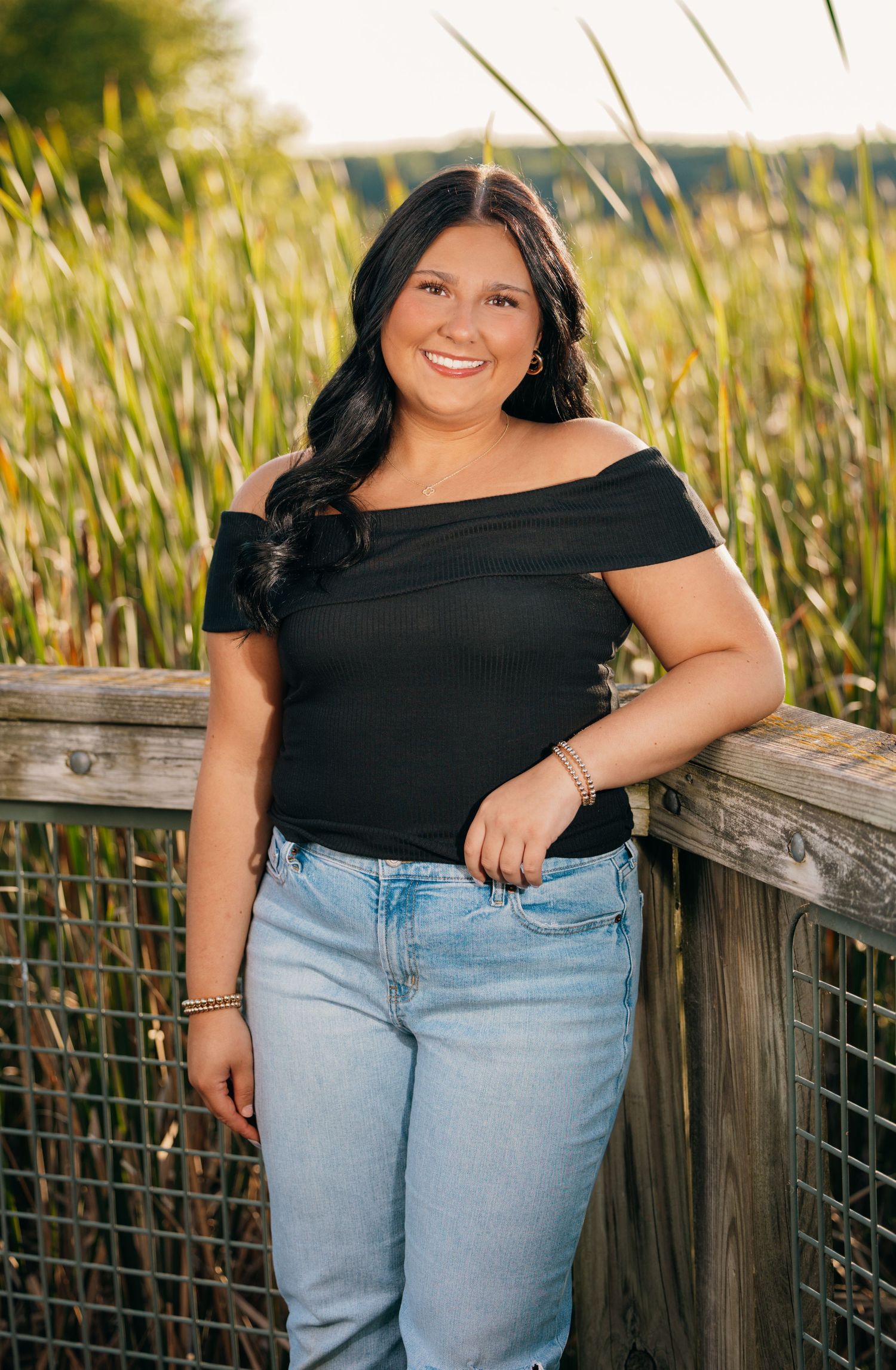 Person in black top and light jeans poses on wooden deck by tall grass.