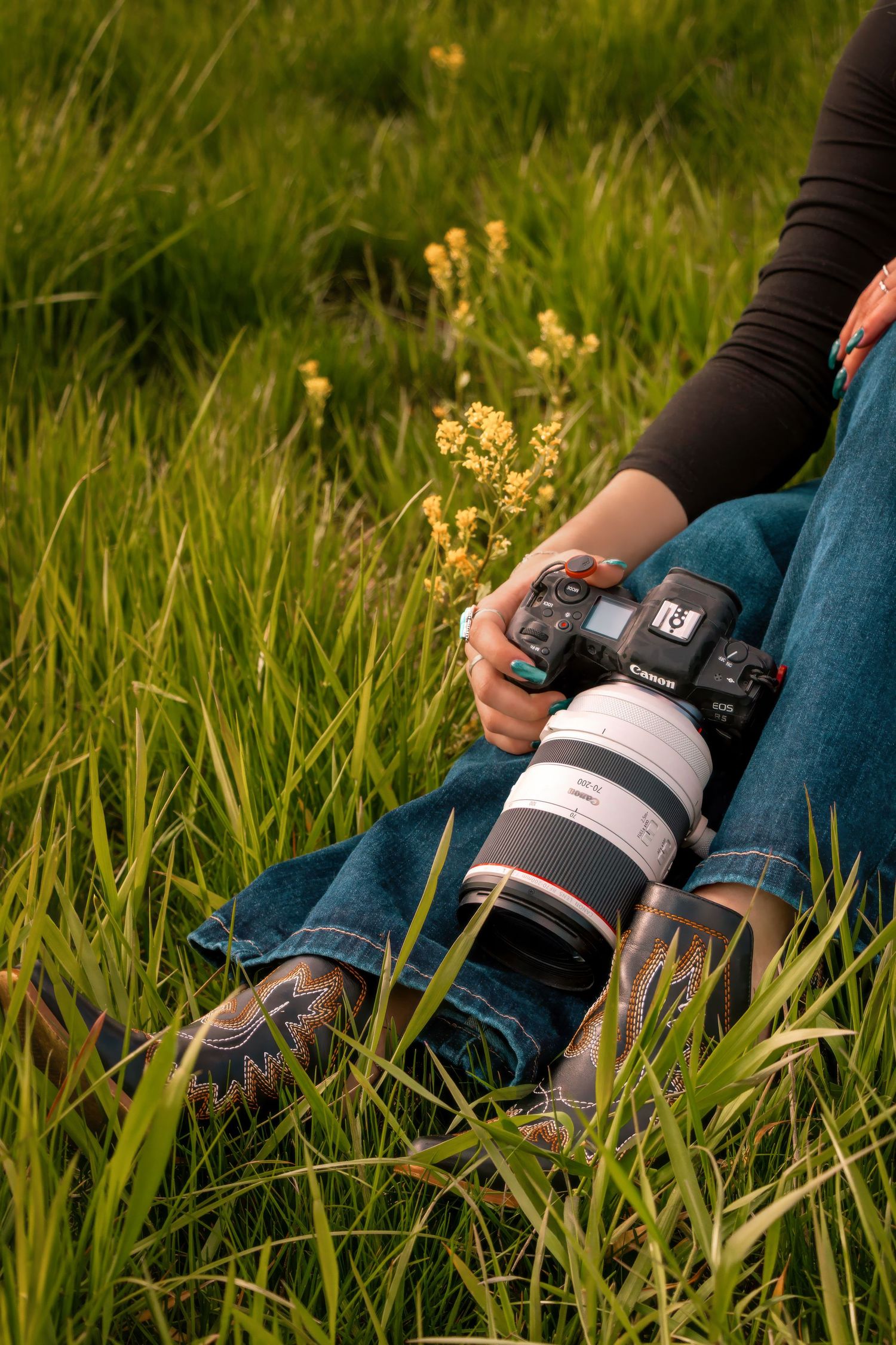Photographer sitting in green meadow holding Canon camera with telephoto lens in golden light.