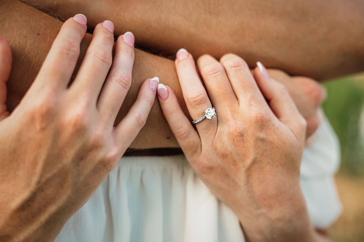 Close-up of hands with an engagement ring intertwined against white fabric.