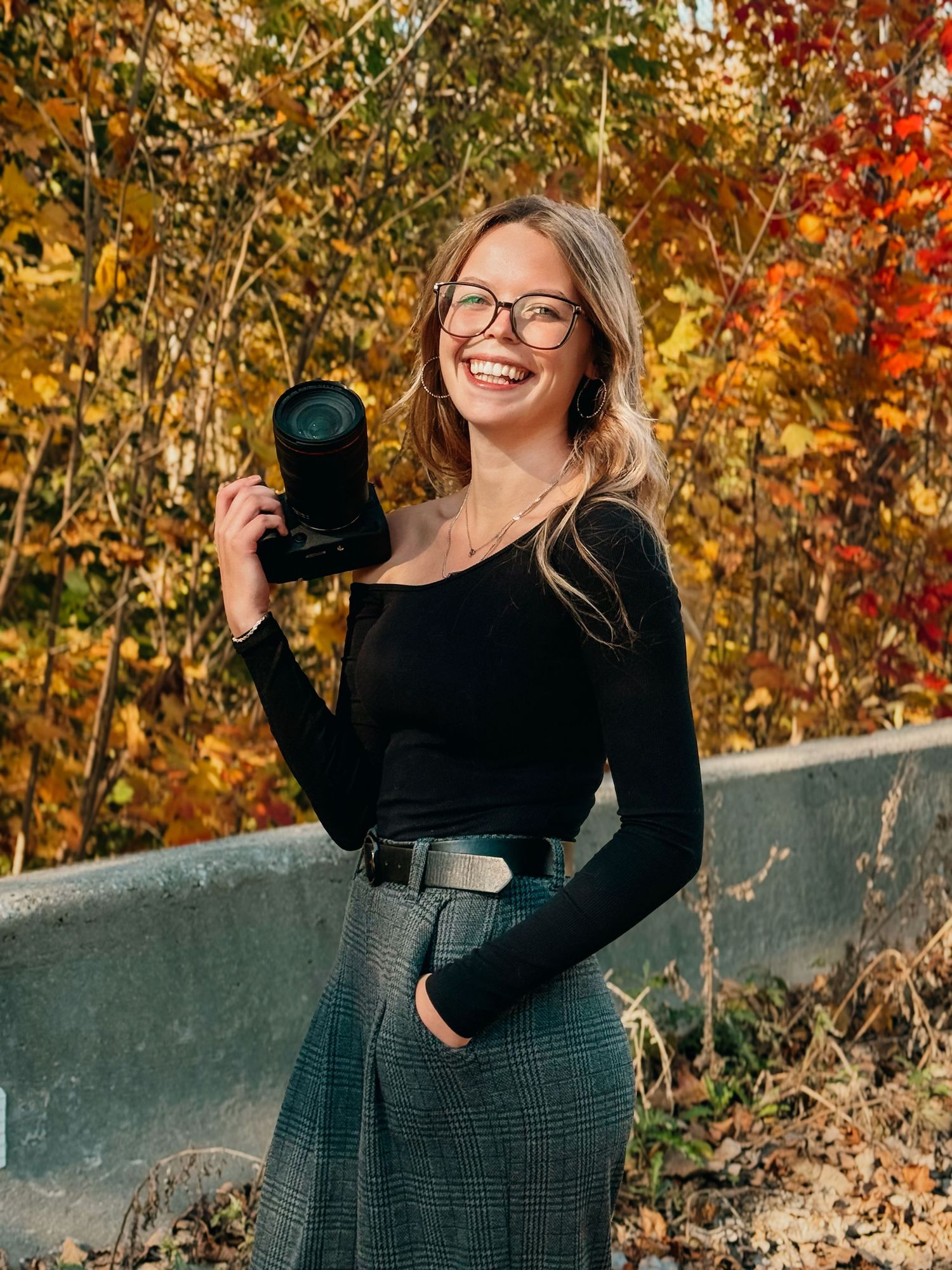 A photographer in a black sweater poses with a camera against vibrant autumn foliage and a stone wall backdrop.
