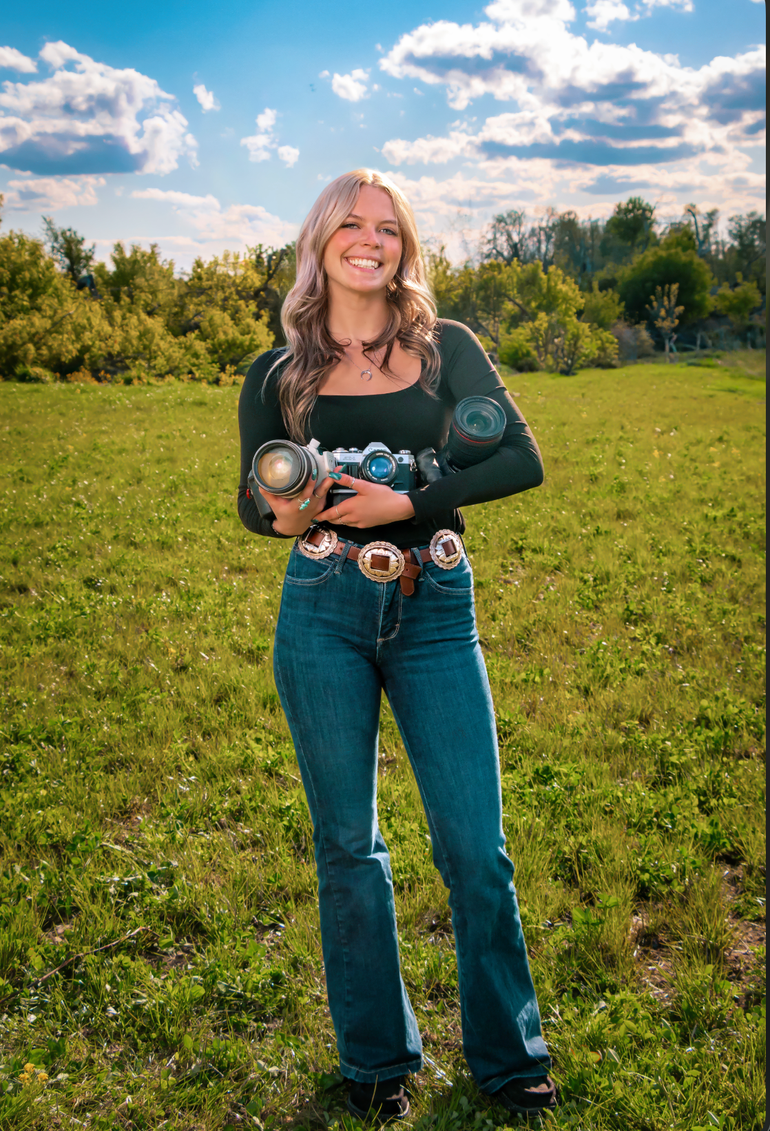Smiling young woman holding cameras standing in a green field wearing jeans and a dark top on a sunny day.