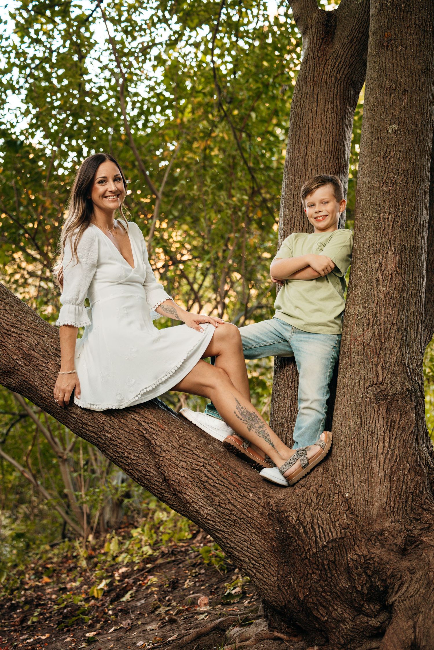A casual outdoor portrait of two people sitting on a large tree branch in a park setting.