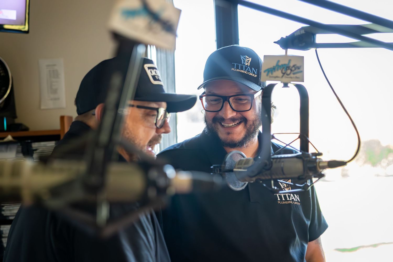 Two people wearing black shirts share a laugh while recording in a podcast studio with microphones.