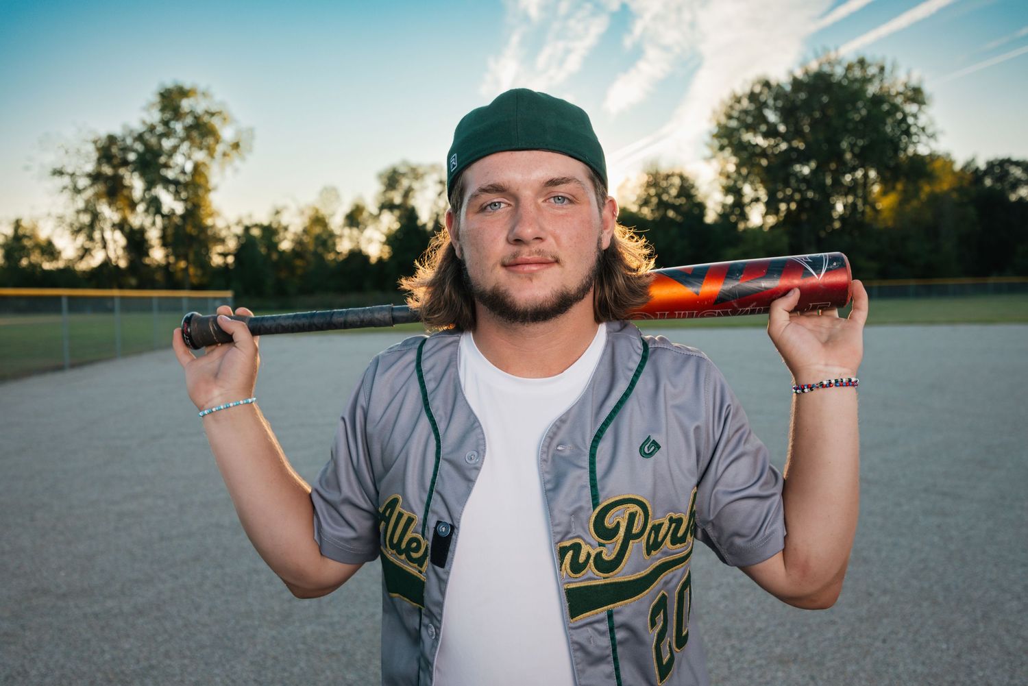 Baseball player wearing green jersey and cap stands in field at sunset.