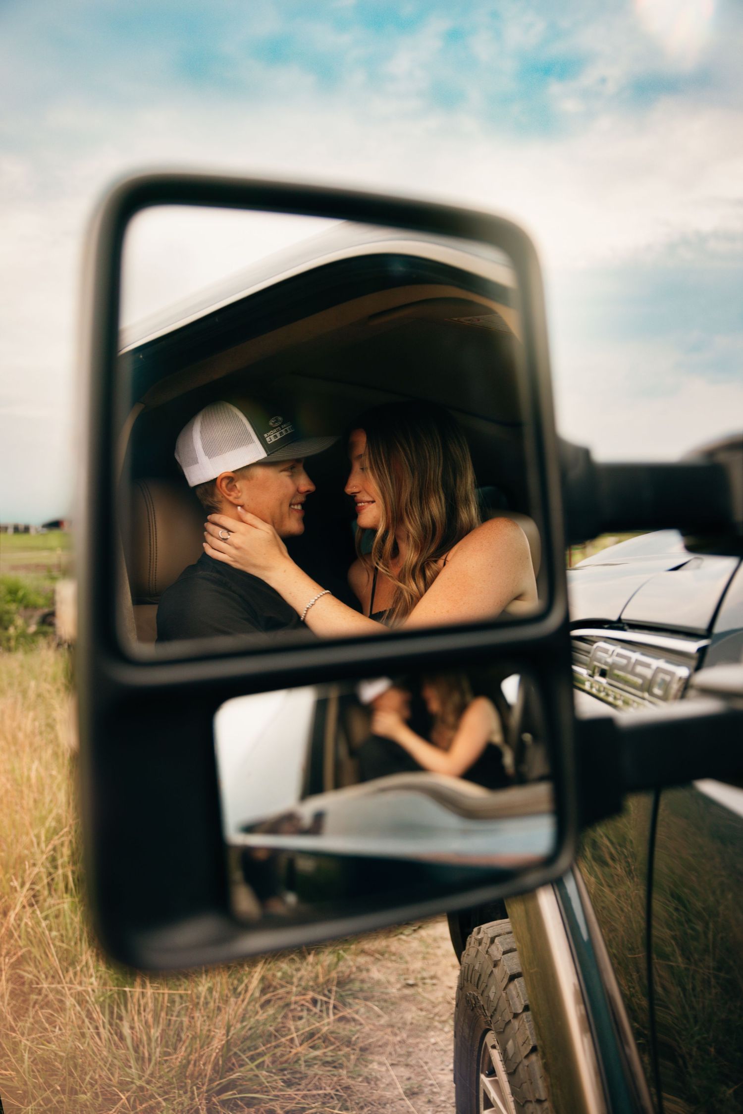 Side mirror reflection captures intimate couple moment in parked car on country road.