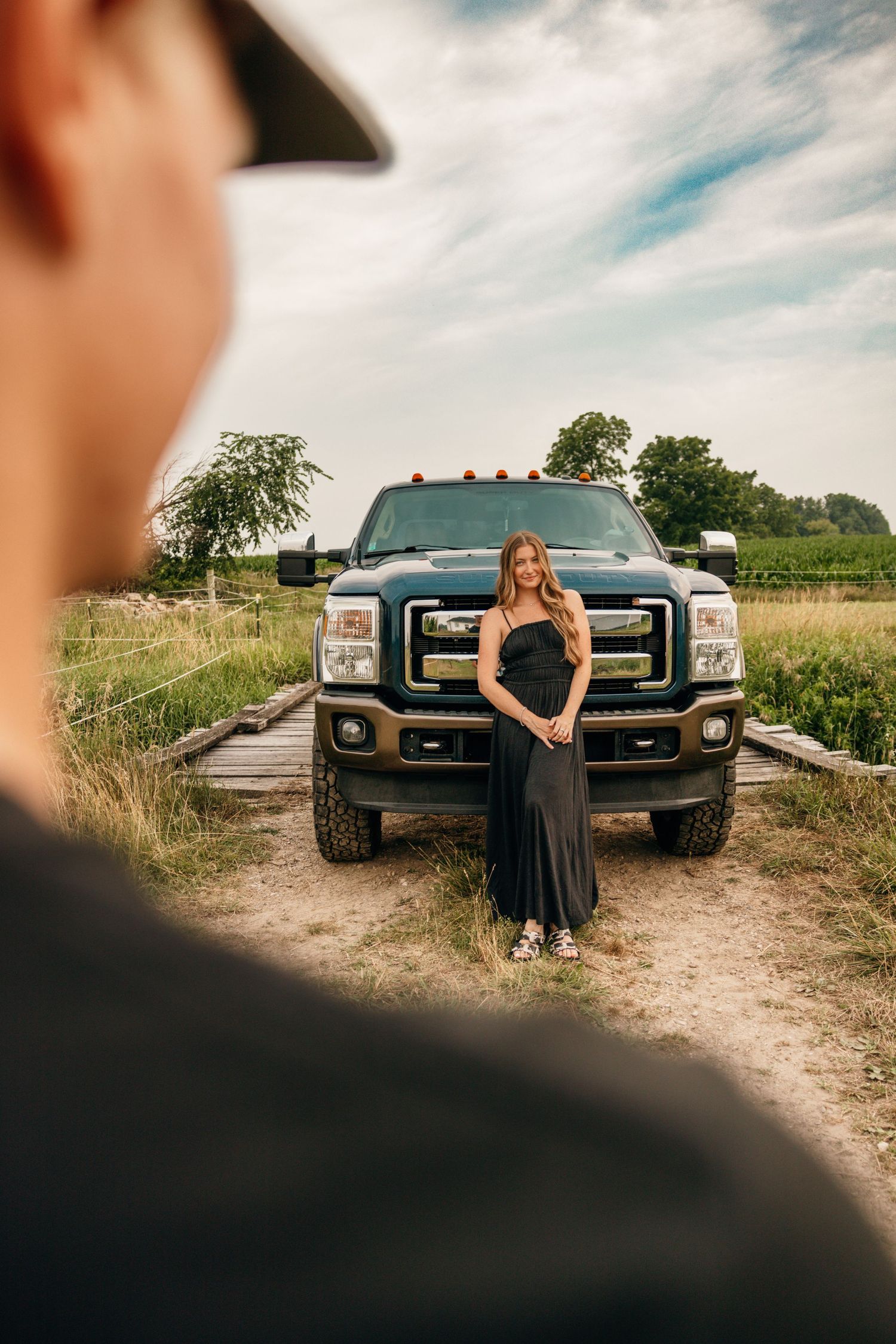Woman in black dress poses in front of large pickup truck on dirt road.