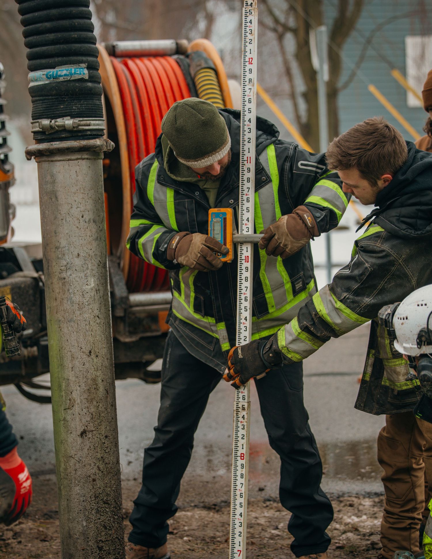 Construction workers in high-visibility gear collaborate on measuring and aligning equipment at a worksite.
