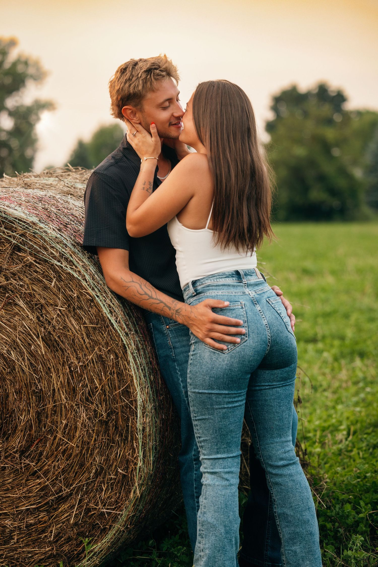 Romantic couple embraces against round hay bale in rural field at sunset.