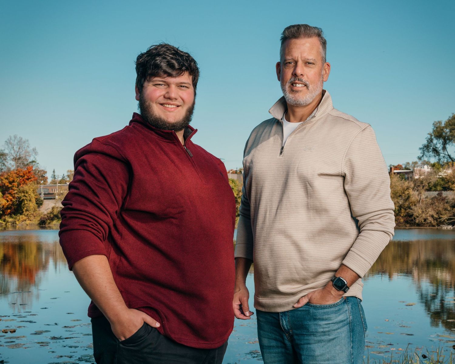 Two people in sweaters pose for a portrait by an autumn lake with fall foliage in background.