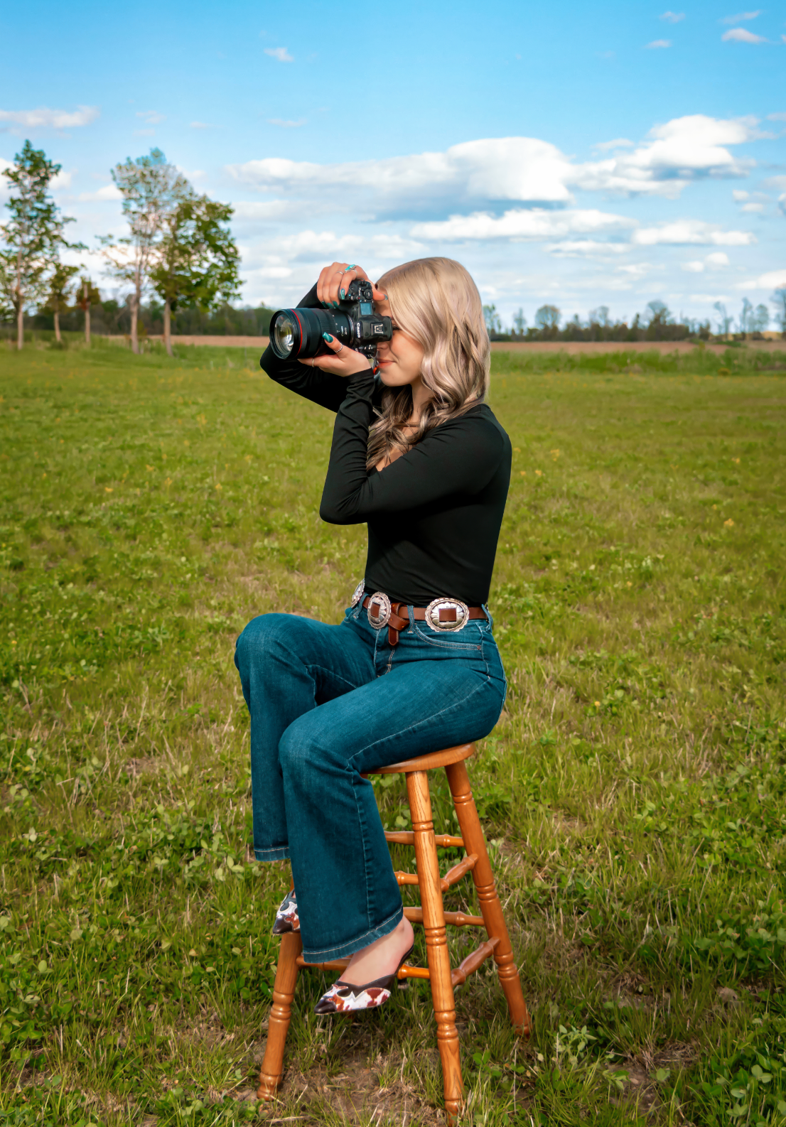 Young blonde woman sitting on a wooden stool in a green field, wearing jeans and black top, photographing with a DSLR camera.