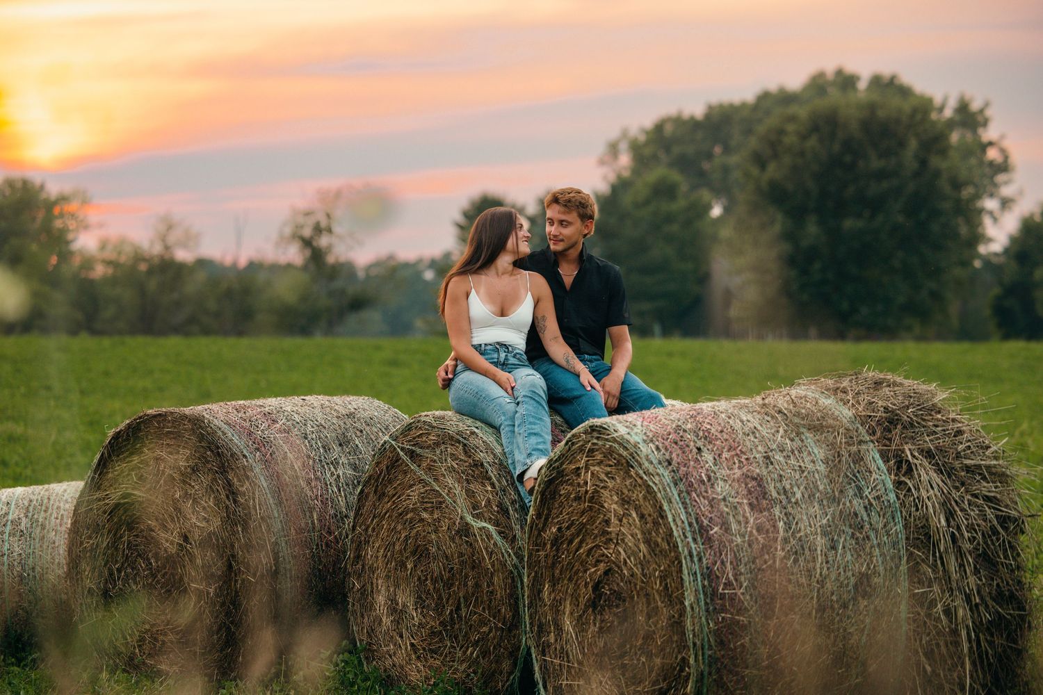 Couple sits on hay bales during sunset in a rural field.