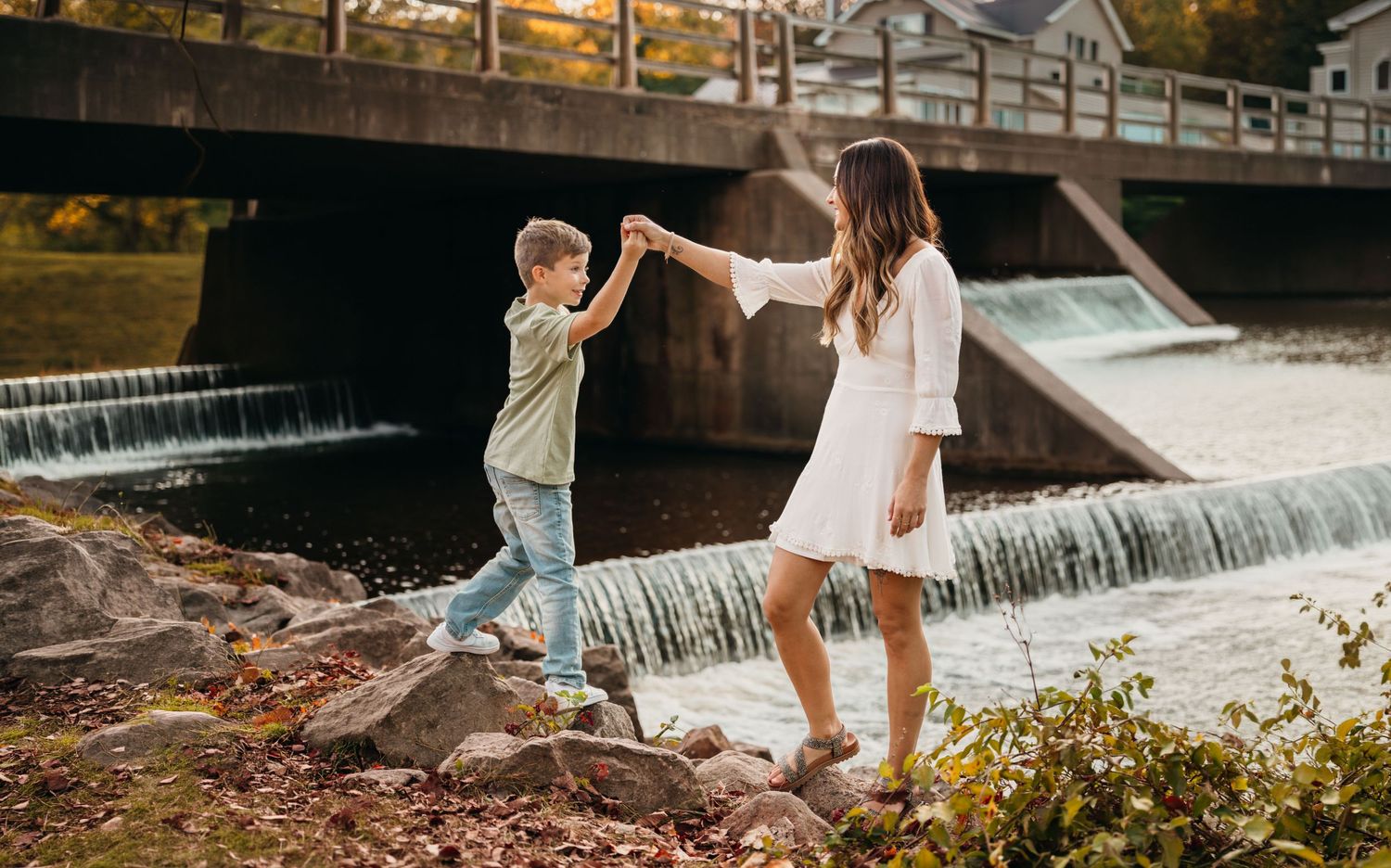 Two people balance and hold hands while standing on rocks by a cascading waterfall.