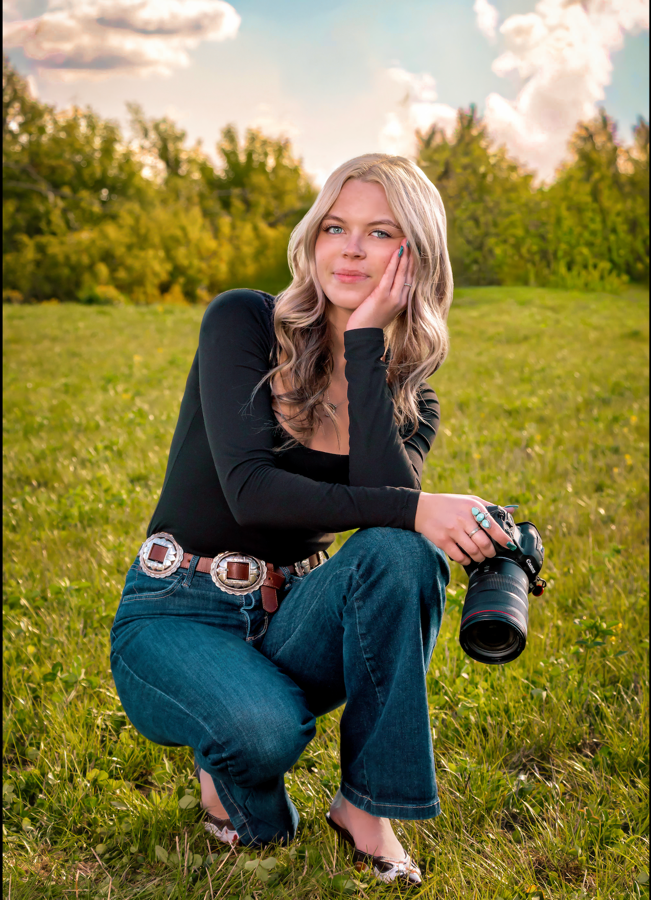 Blonde woman crouching in green field holding DSLR camera, wearing black top, jeans, and decorative belt.