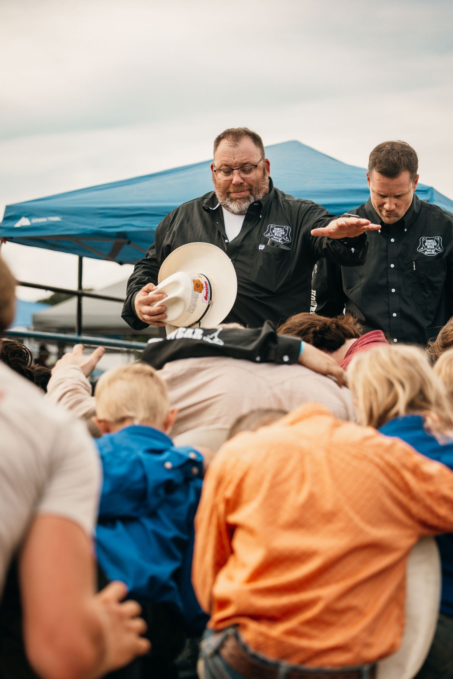 Group huddles under blue tent at outdoor event wearing cowboy hats.