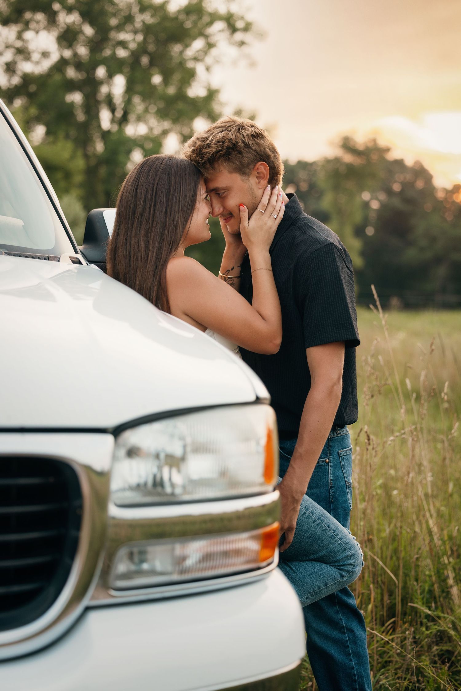 Couple shares intimate moment near white car in tall grass field during sunset.