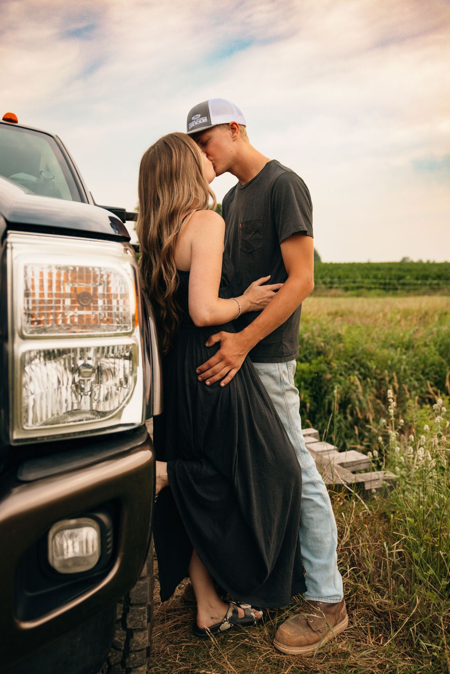 Romantic couple embracing against a pickup truck in a rural field at sunset.
