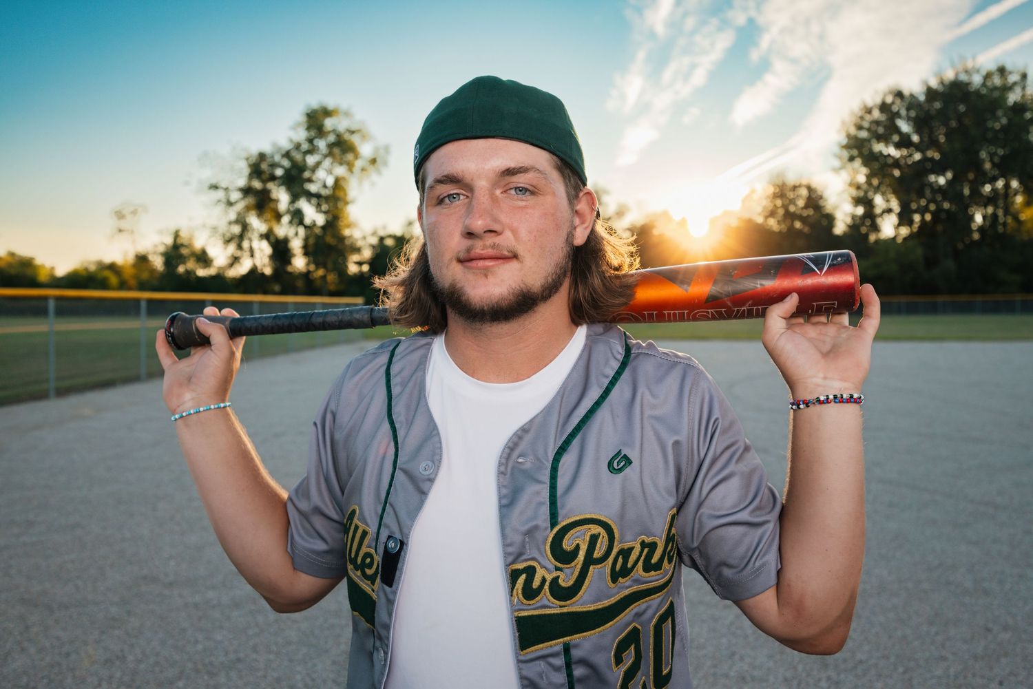 Baseball player in grey jersey and cap poses at sunset on a sports field.