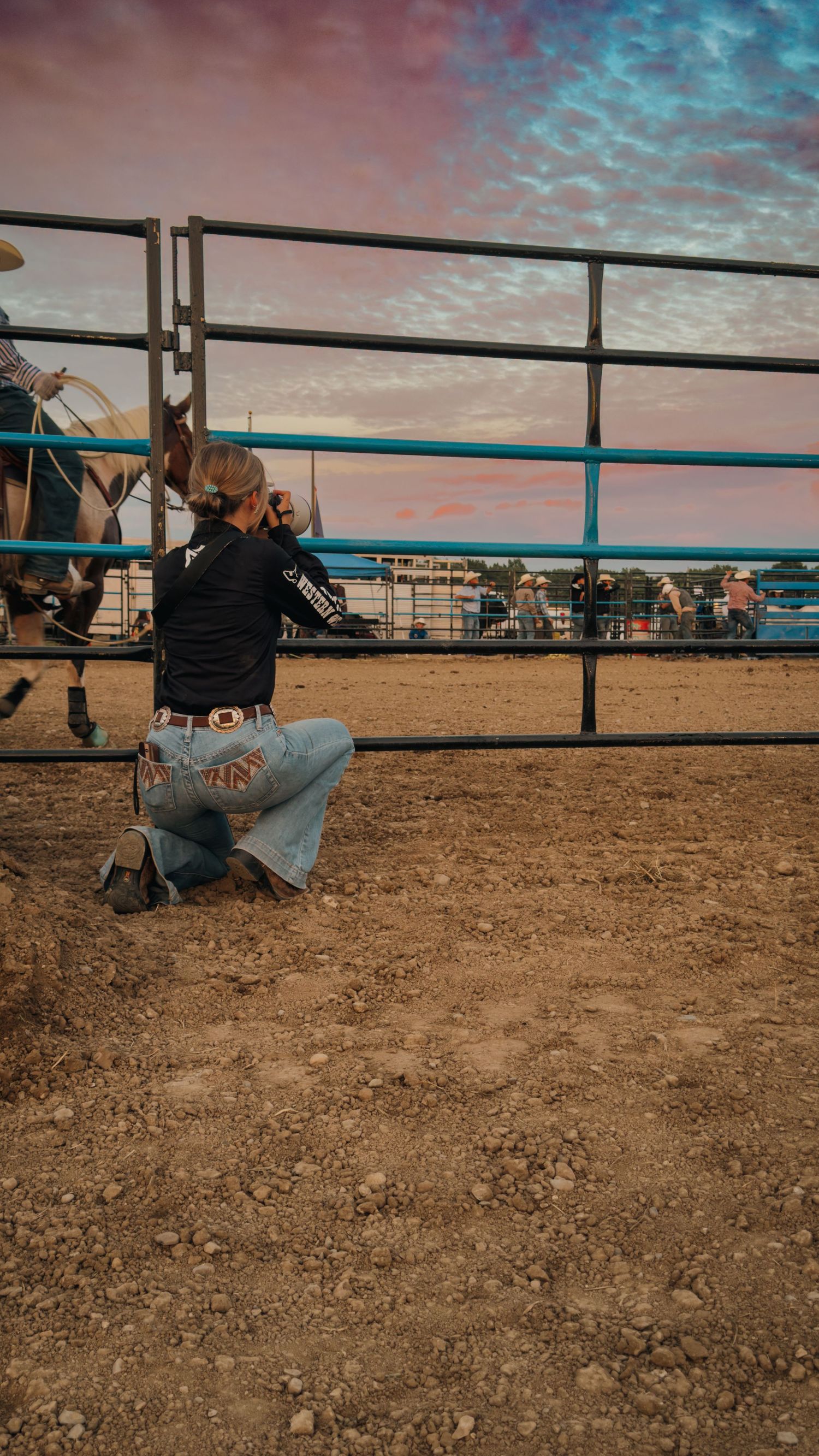 A photographer kneels to capture photos at a horse arena during sunset.