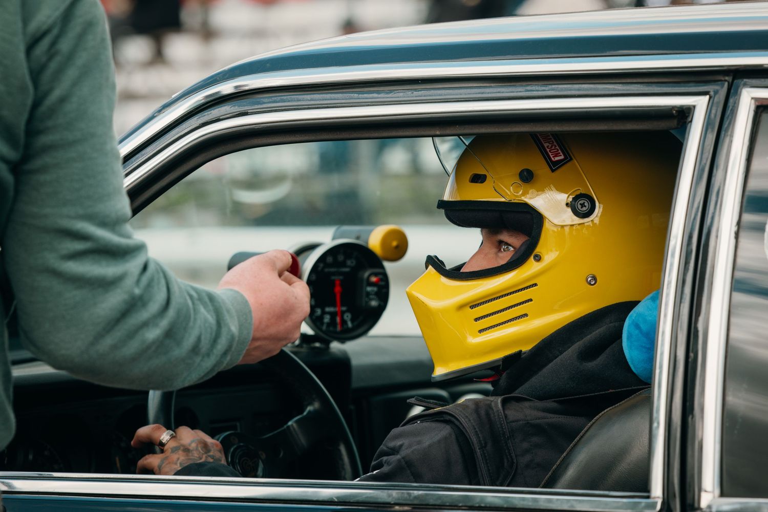 A person wearing a yellow racing helmet sits in a vintage car while being photographed.