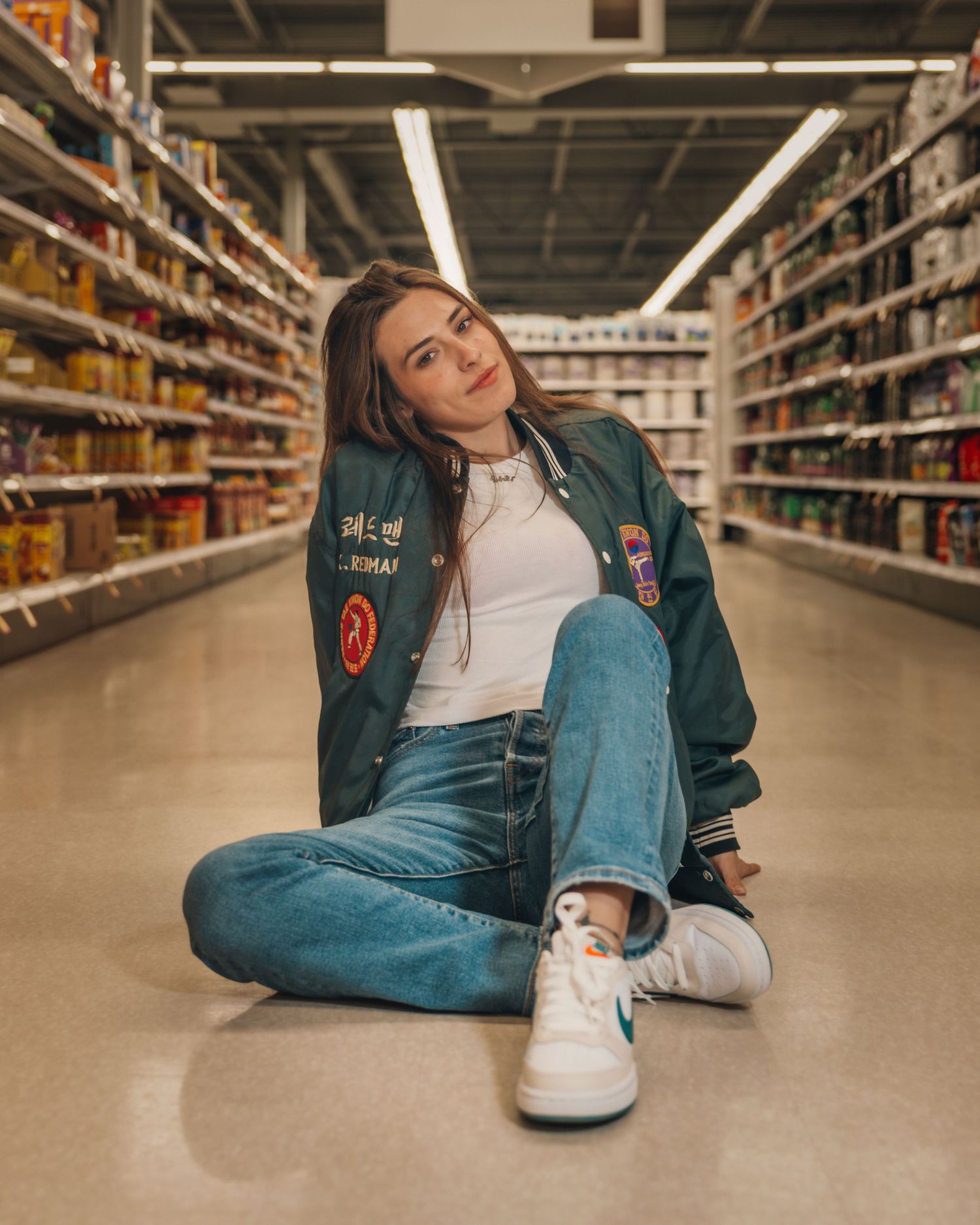 Person relaxing on supermarket floor wearing varsity jacket and jeans.
