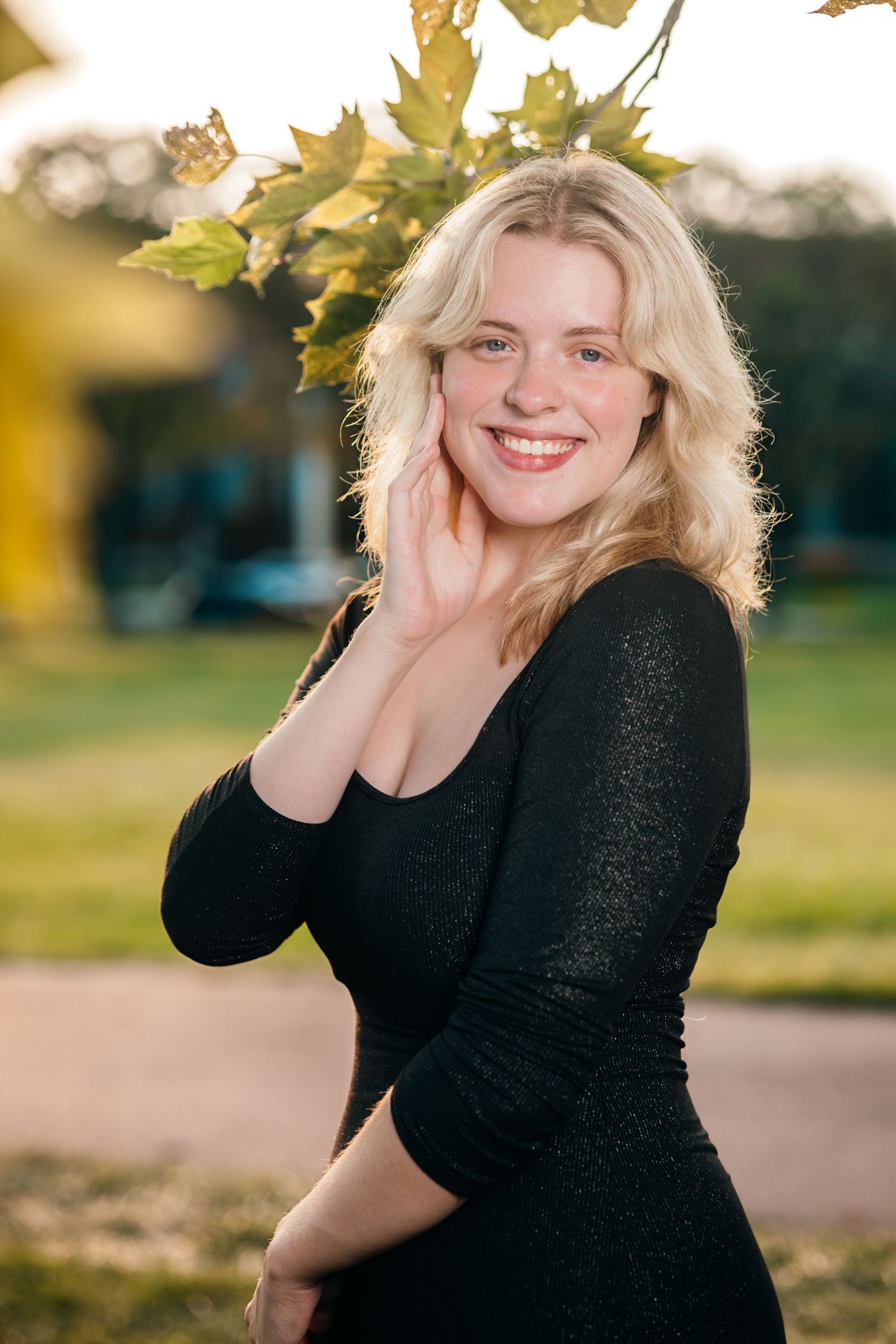 Person in black sweater poses playfully outdoors with autumn leaves in background during golden hour.