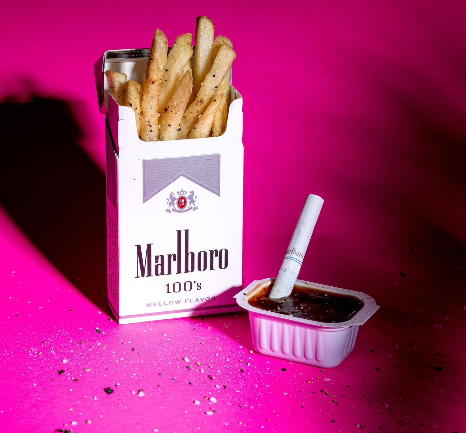 Pack of Marlboro cigarettes against vibrant pink background with scattered tobacco.