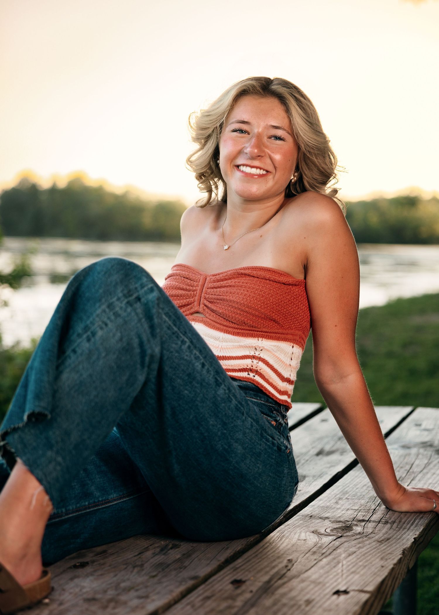 Person in coral top and jeans relaxes by waterfront at sunset.