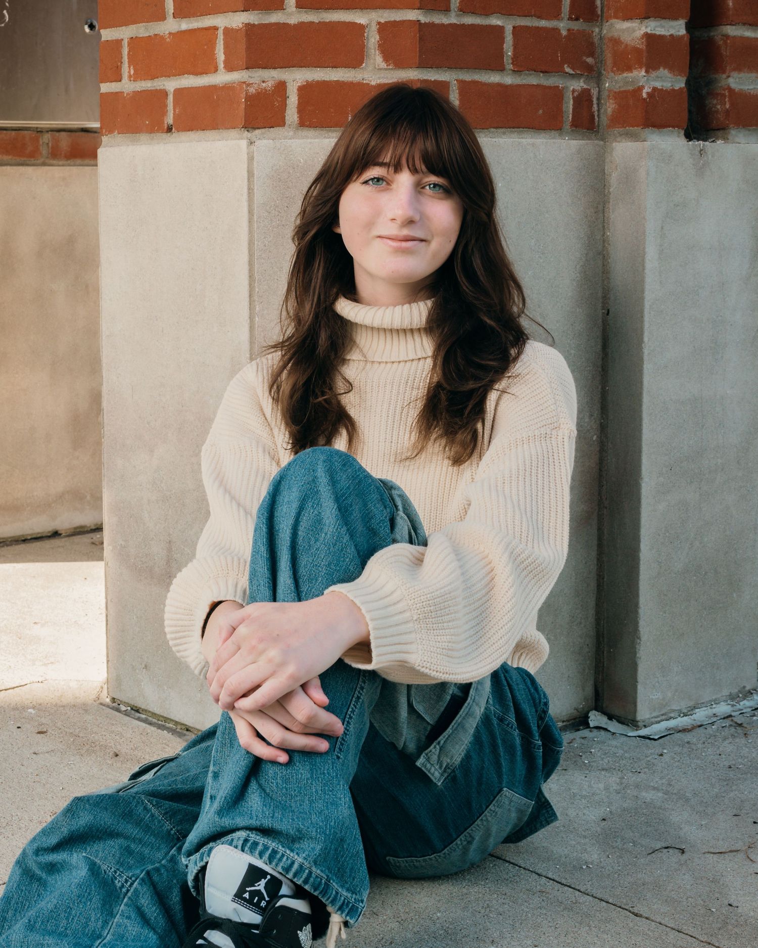 Casual portrait sitting against brick wall wearing beige sweater and denim.