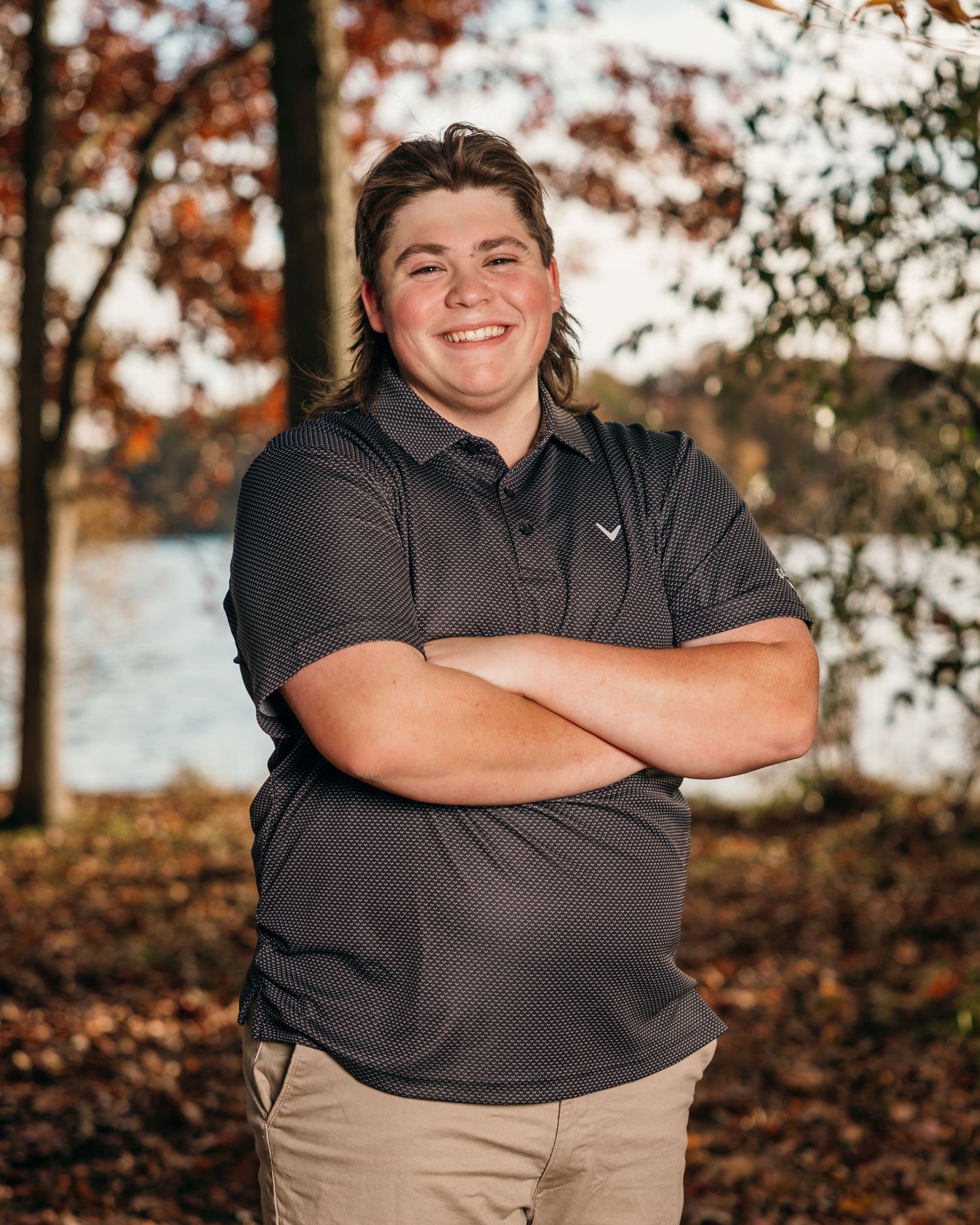 Person in dark polo shirt and khaki pants poses outdoors near autumn trees.