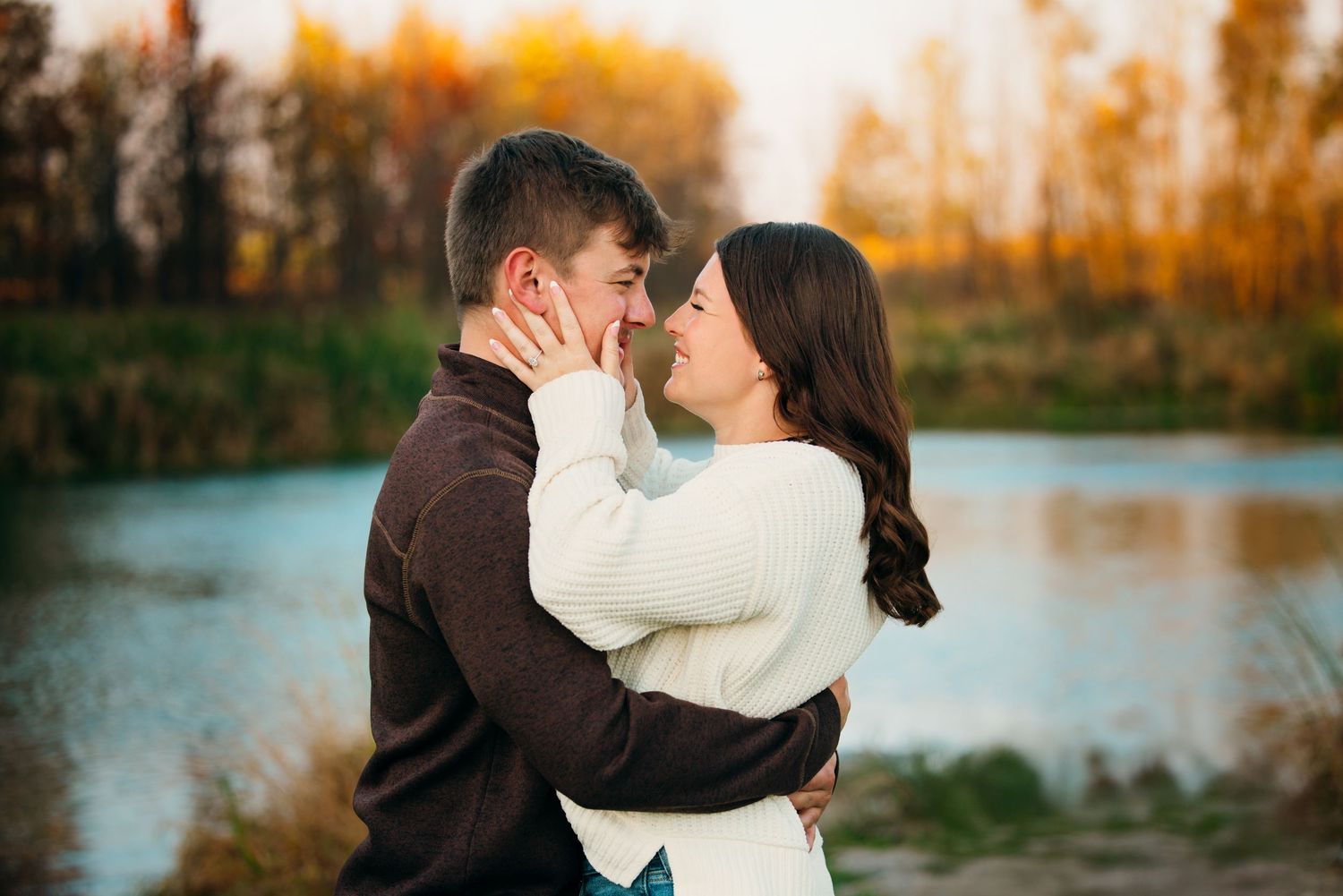 Couple in white and brown sweaters embrace by a lake during autumn sunset.