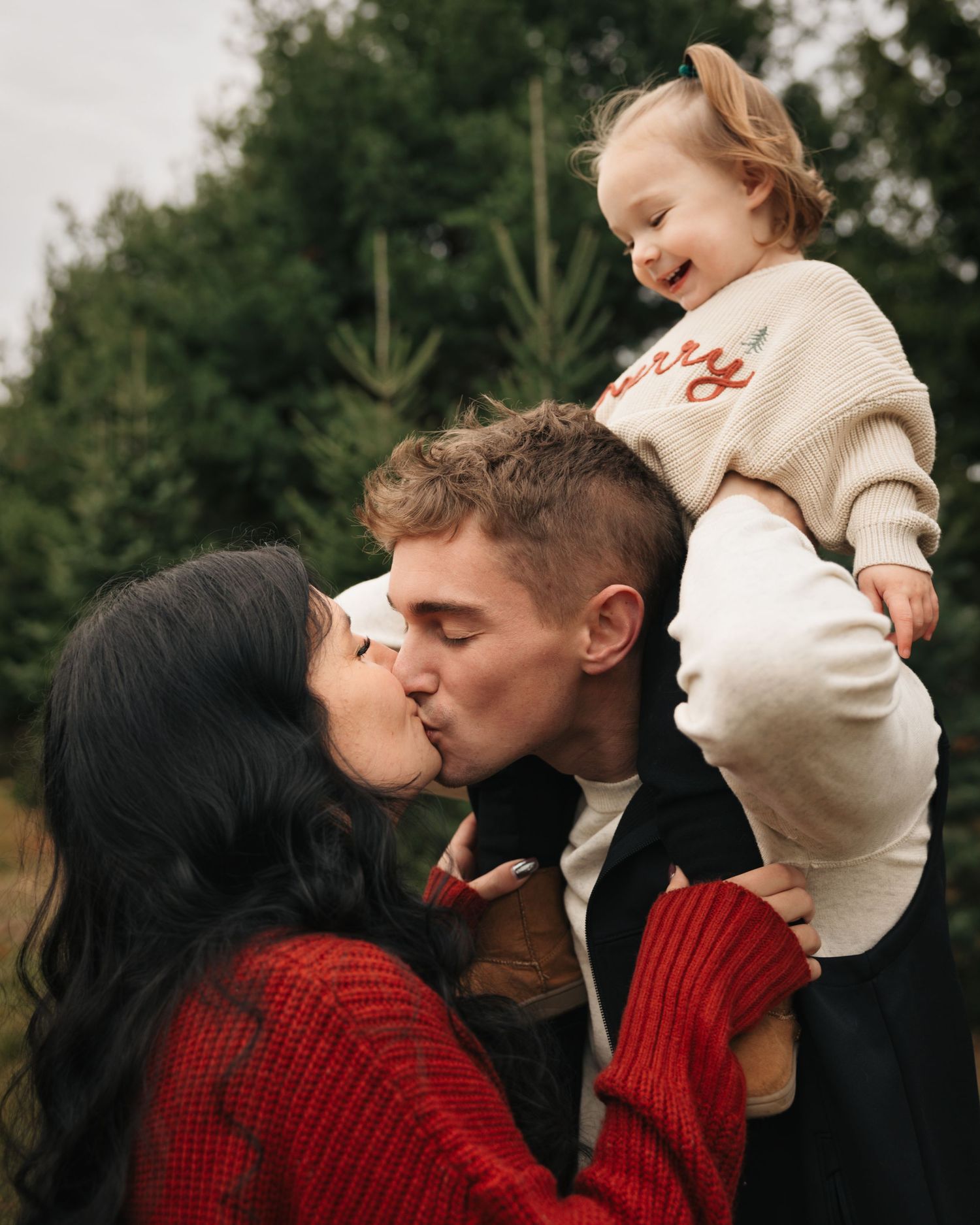 Family sharing a tender moment at a Christmas tree farm while wearing cozy winter sweaters.