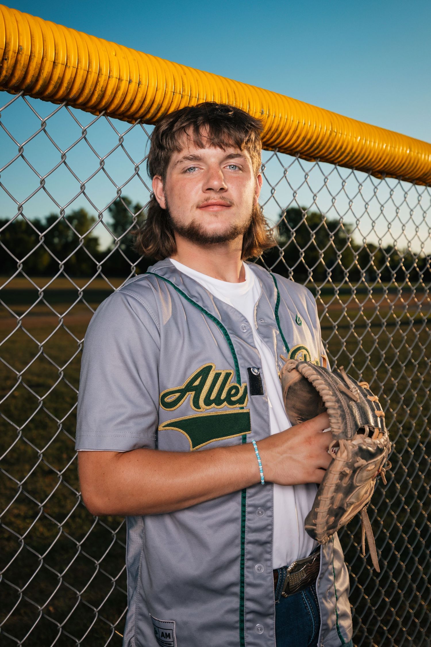 Baseball player in Allen Park uniform poses against chain link fence with yellow padding.