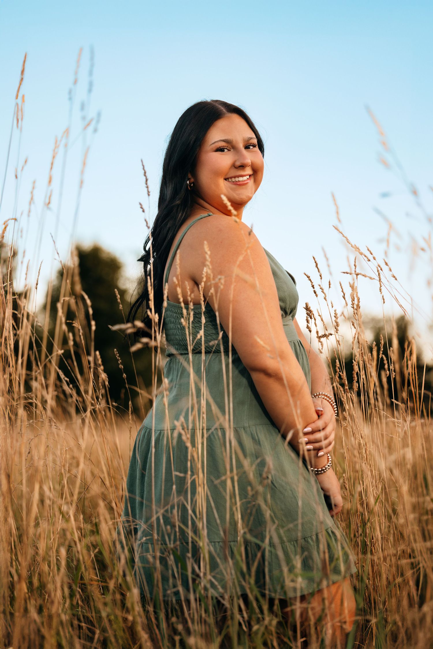 A woman in a teal dress poses in a field of tall grass during golden hour.