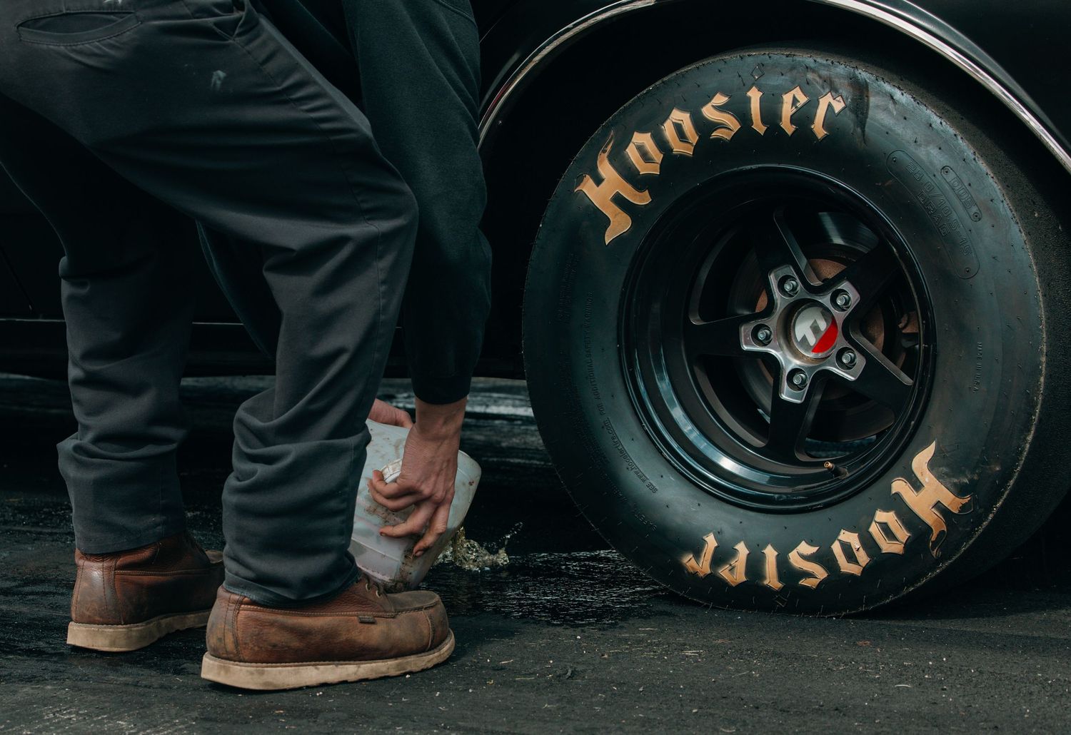 Close-up of Hoosier racing tires and boots on black asphalt surface.