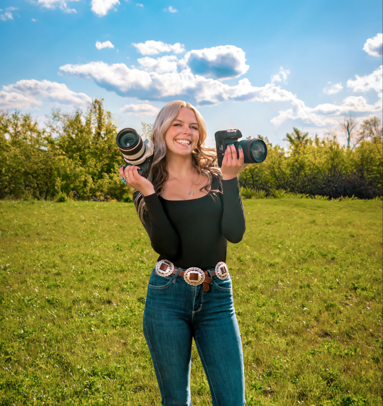 Young blonde woman smiling outdoors holding two professional cameras in a green field with blue sky.