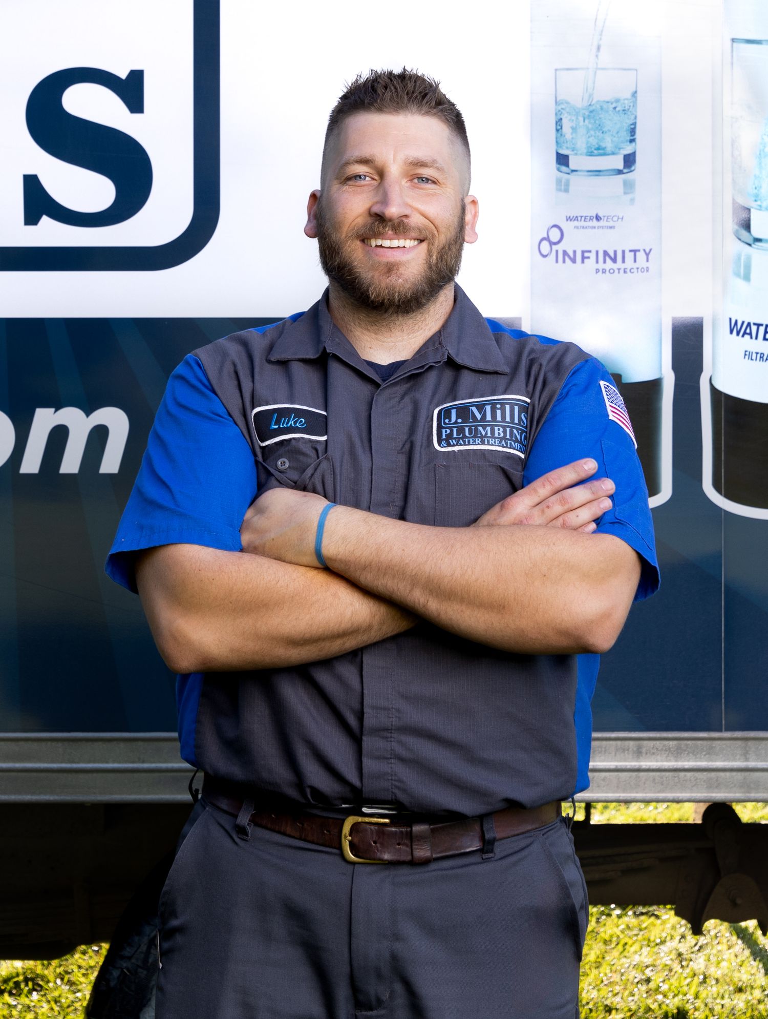 Service technician in grey vest and blue shirt poses with arms crossed by company vehicle.